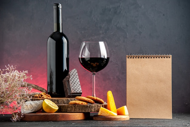 Front view black wine bottle red wine in glass cheese cut lemon pieces of dark chocolate biscuits on wooden boards notebook on red table