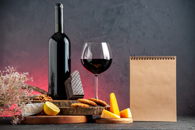 Front view black wine bottle red wine in glass cheese cut lemon pieces of dark chocolate biscuits on wooden boards notebook on red table