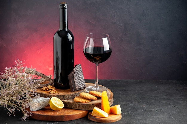 Front view black wine bottle red wine in glass cheese cut lemon pieces of dark chocolate on wooden boards dried flower branch on red table copy place