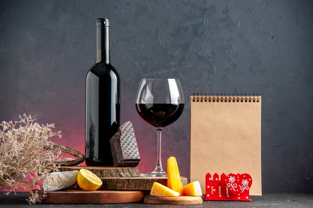 正面図ガラスチーズカットレモンの黒ワインボトルワイン木の板にダークチョコレートのかけら赤いテーブルにドライフラワーブランチメモ帳