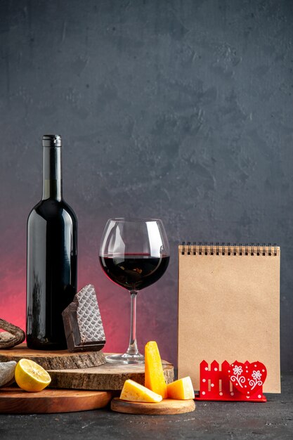 Вид спереди бутылка черного вина, вино в стеклянном сыре, нарезанные кусочками лимона темного шоколада на деревянных досках на красном столе