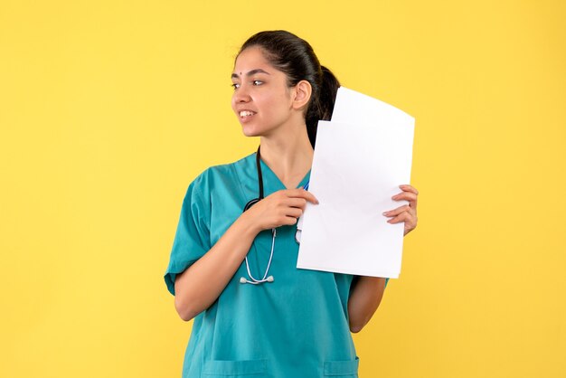 Front view blessed pretty female doctor holding papers standing on yellow background