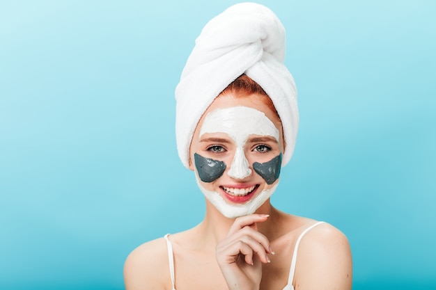 Free photo front view of blissful caucasian woman with face mask. studio shot of pleasant girl with towel on head posing on blue background.