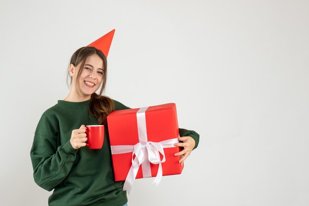 Front view blissful girl with party cap holding her xmas gift and cup