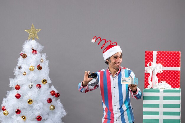 Front view blissful man holding card and travel ticket around xmas tree and presents