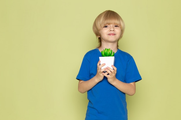 A front view blodne smiling boy in blue t-shirt holding little green plant on the stone colored space