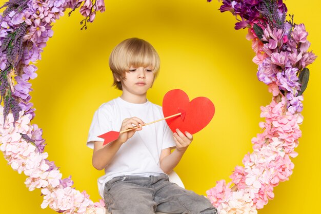 A front view blonde boy cute adorable in white t-shirt holding heart shape on the flower made desk on the yellow floor