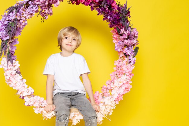 A front view blonde boy sweet cute adorable in white t-shirt sitting on the flower made stand on the yellow desk