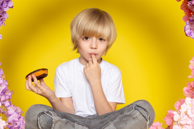 A front view blonde boy in white t-shirt eating choco donuts sitting on the flower made stand on the yellow space