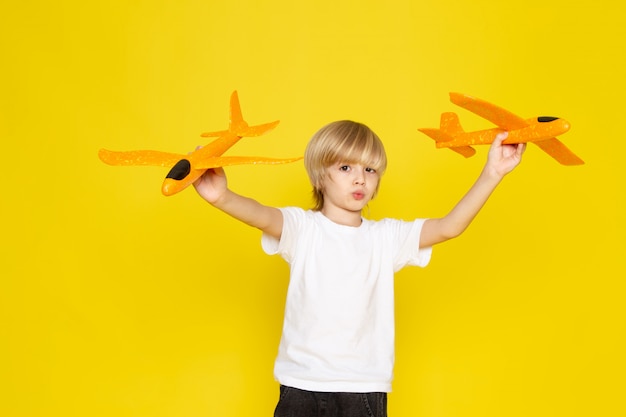 Front view blonde boy in white t-shirt playing with toy orange planes on the yellow floor