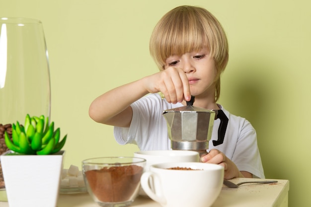 A front view blonde boy in white t-shirt preparing coffee on the stone colored wall