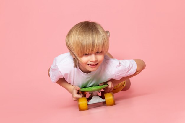 Вид спереди белокурый мальчик в белой футболке катается на скейтборде на розовом полу