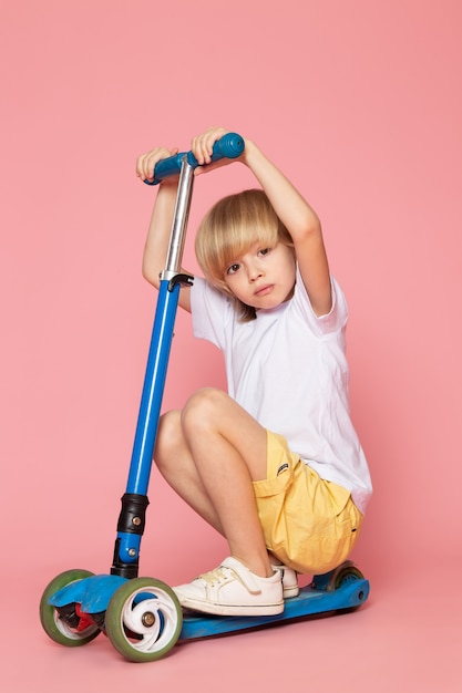 A front view blonde boy in white t-shirt and yellow jeans riding blue scooter on the pink floor