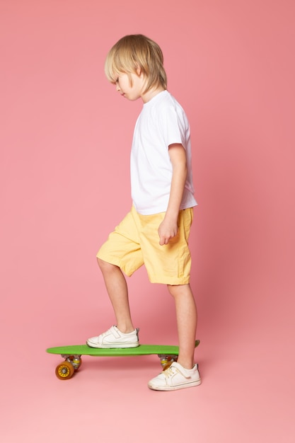 Free photo a front view blonde boy in white t-shirt and yellow jeans riding green skateboard on the pink space