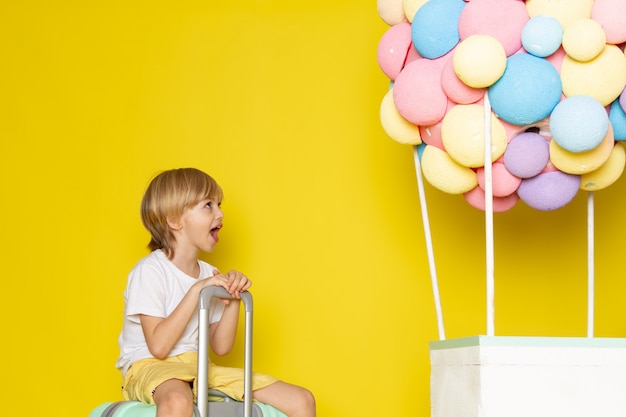 front view blonde boy in white t-shirt and yellow shorts along with colorful balloons on the yellow