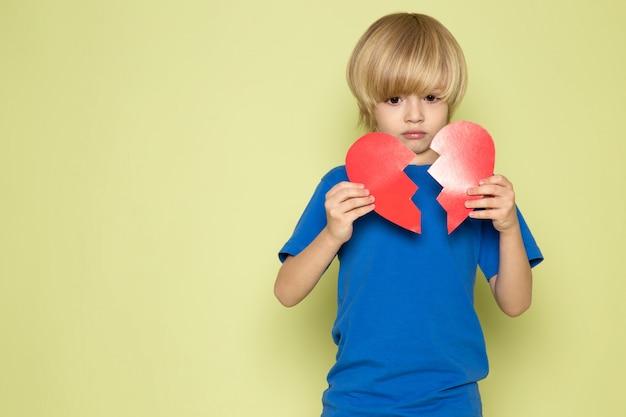 A front view blonde cute boy in blue t-shirt tearing heart shape on the stone colored space