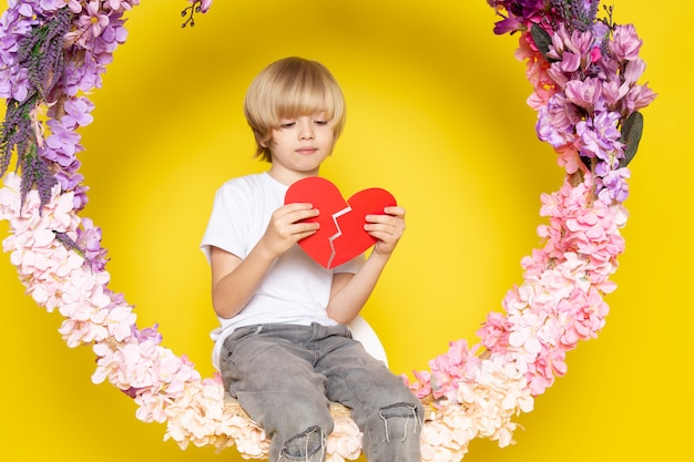 Free photo a front view blonde cute boy in white t-shirt holding heart shape sitting on the flower made stand on the yellow floor