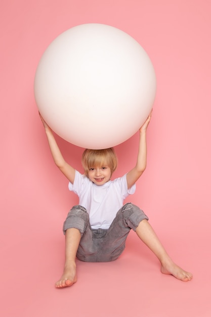 ピンクの机の上の丸い白いボールで遊ぶ白いtシャツで正面金髪のかわいい男の子