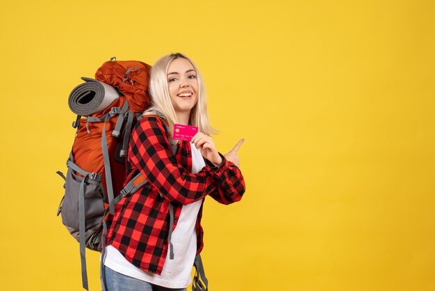 Front view blonde girl with her backpack holding card pointing at behind