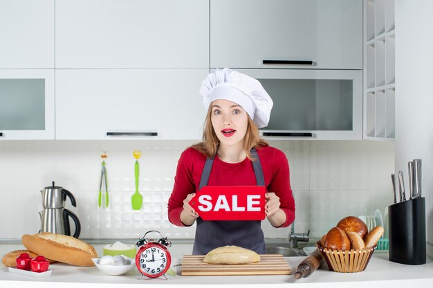 Front view blonde pretty woman in cook hat and apron holding sale sign in the kitchen