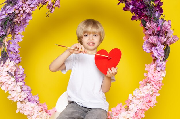 A front view blonde smiling boy in white t-shirt holding heart shape on the yellow floor