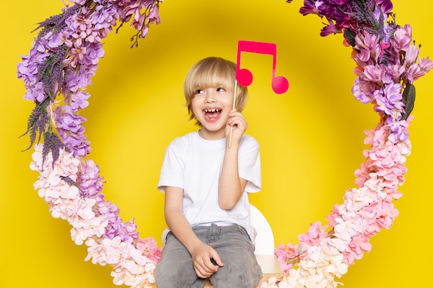 A front view blonde smiling boy in white t-shirt holding pink note on the flower made stand on the yellow desk