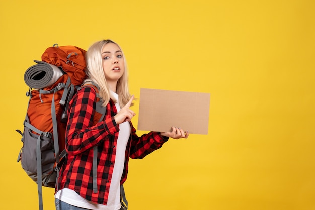 Front view blonde traveler woman with her backpack holding cardboard standing on yellow wall