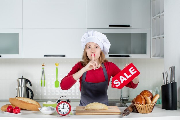 Foto gratuita vista frontale donna bionda con cappello da cuoco e grembiule con cartello di vendita che fa cantare il silenzio in cucina