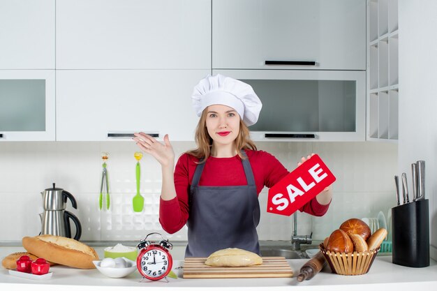 Foto gratuita vista frontale giovane donna bionda in cappello da cuoco e grembiule con cartello di vendita in cucina