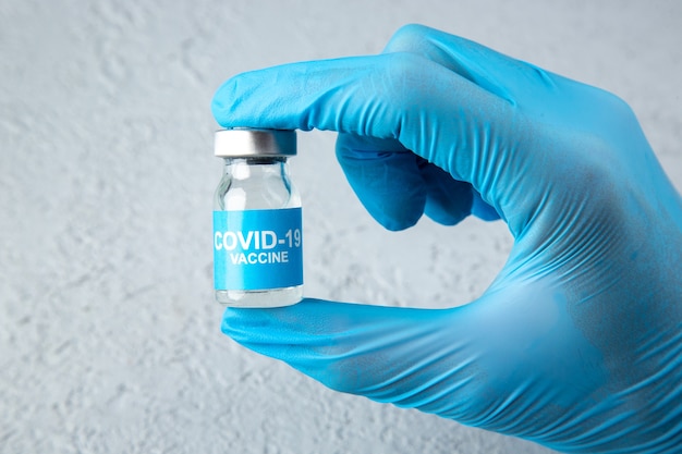 회색 모래 배경에 covid-백신이 있는 닫힌 앰플을 들고 있는 파란색 장갑의 전면 보기