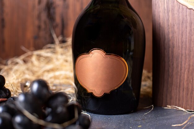 A front view bottle of alcohol black bottle with golden cap along with black grapes and green leaves on the brown background drink winery alcohol