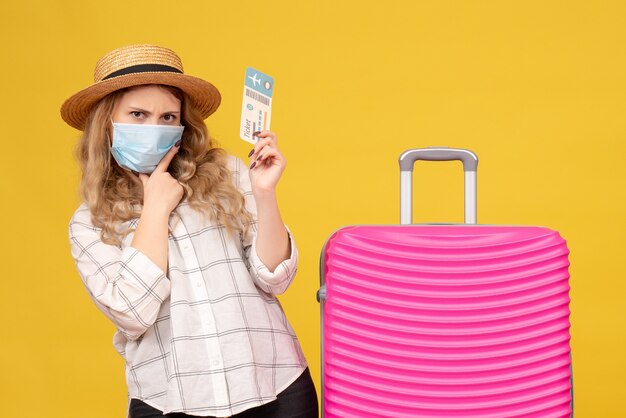 Front view of brainstorming travelling girl wearing mask showing ticket and standing near her pink bag on yellow 