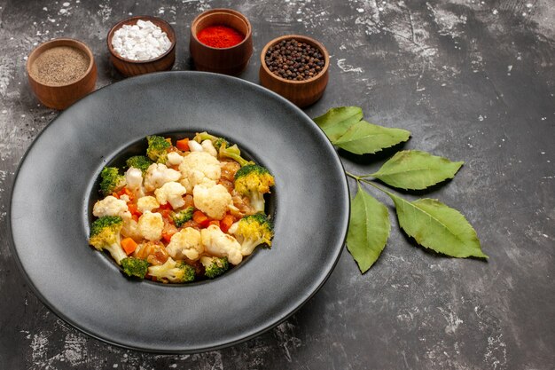 Front view broccoli and cauliflower salad in black bowl different spices in bowls leaves on dark background