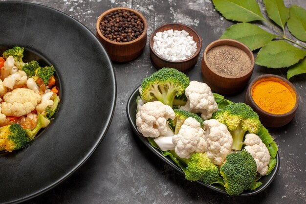 Front view broccoli and cauliflower salad in black bowl raw broccoli and cauliflower on plate different spices on dark background
