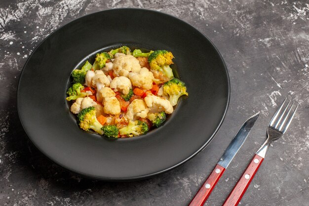 Front view broccoli and cauliflower salad on black oval plate fork and knife on dark background