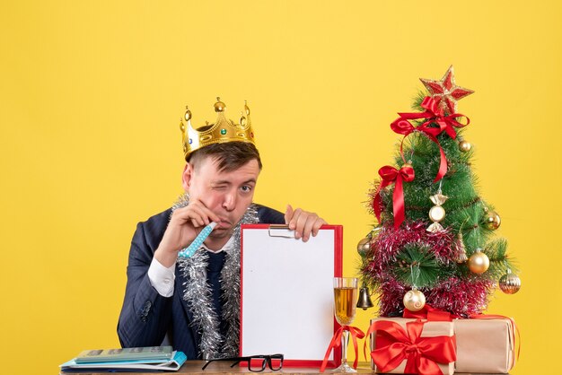 Vista frontale dell'uomo di affari che lampeggia alla macchina fotografica che si siede al tavolo vicino all'albero di natale e regali sulla parete gialla