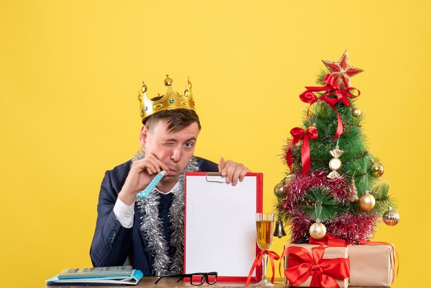 Вид спереди делового человека, моргающего в камеру, сидящего за столом возле рождественской елки и подарков на желтой стене