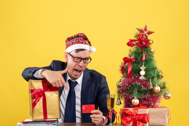 Вид спереди делового человека, моргающего глазами, сидящего за столом возле рождественской елки и подарков на желтой стене