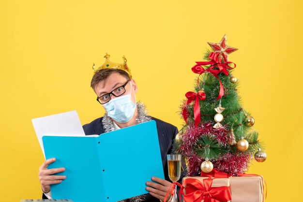 Front view of business man checking documents sitting at the table near xmas tree and presents on yellow wall