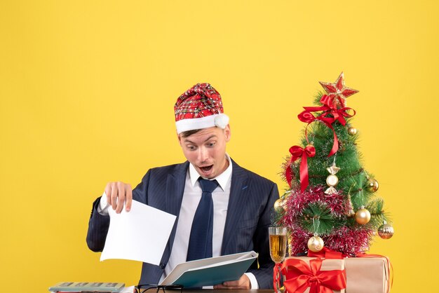 Front view of business man checking papers sitting at the table near xmas tree and presents on yellow wall