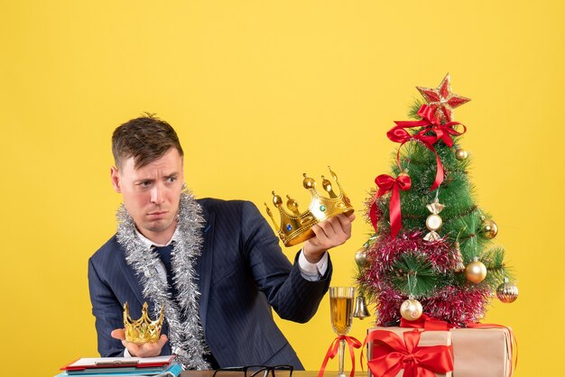 Front view business man comparing his crowns sitting at the table near xmas tree and presents on yellow background