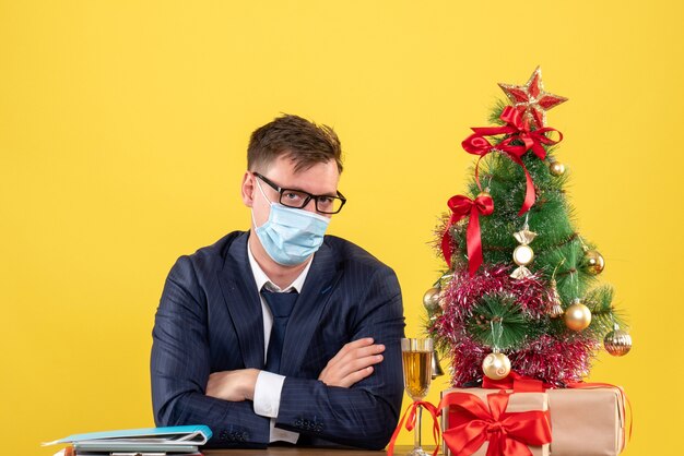 Front view business man crossing hands sitting at the table near xmas tree and presents on yellow background