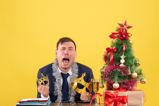 Front view of business man crying while sitting at the table near xmas tree and presents on yellow wall