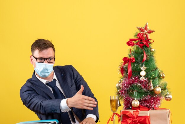 Vista frontale dell'uomo di affari che dà la mano che si siede al tavolo vicino all'albero di natale e presenta sulla parete gialla