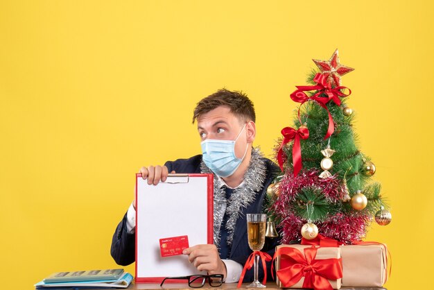 Front view of business man holding clipboard and credit card sitting at the table near xmas tree and presents on yellow