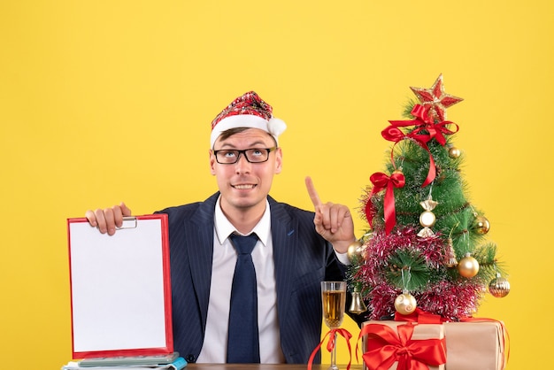 Front view of business man holding clipboard looking up sitting at the table near xmas tree and presents on yellow