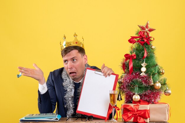 Front view of business man holding clipboard and noisemaker sitting at the table near xmas tree and presents on yellow
