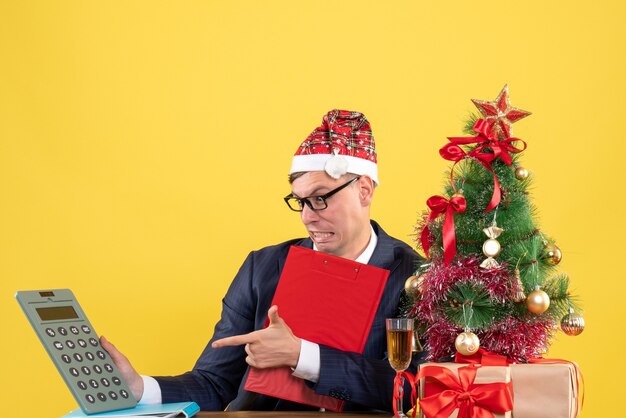 Front view of business man holding clipboard sitting at the table near xmas tree and presents on yellow