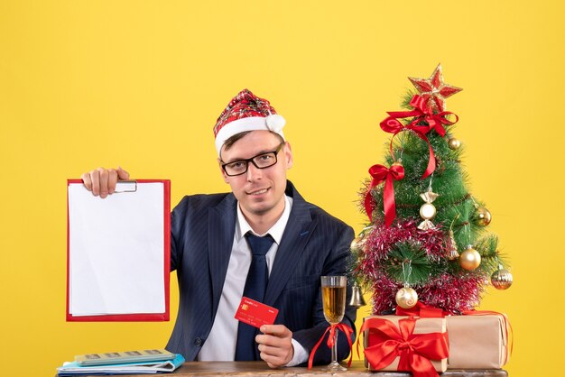 Front view of business man holding credit card and clipboard sitting at the table near xmas tree and presents on yellow