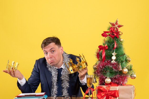 クリスマスツリーの近くのテーブルに座って、黄色でプレゼント両手で王冠を保持しているビジネスマンの正面図